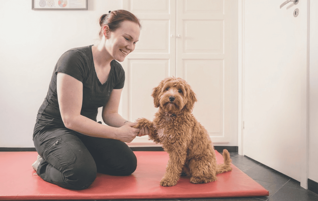 Trine Ahlmann Hansen knæler på en rød måtte og smiler til en fluffy brun hund, mens de holder poter sammen.